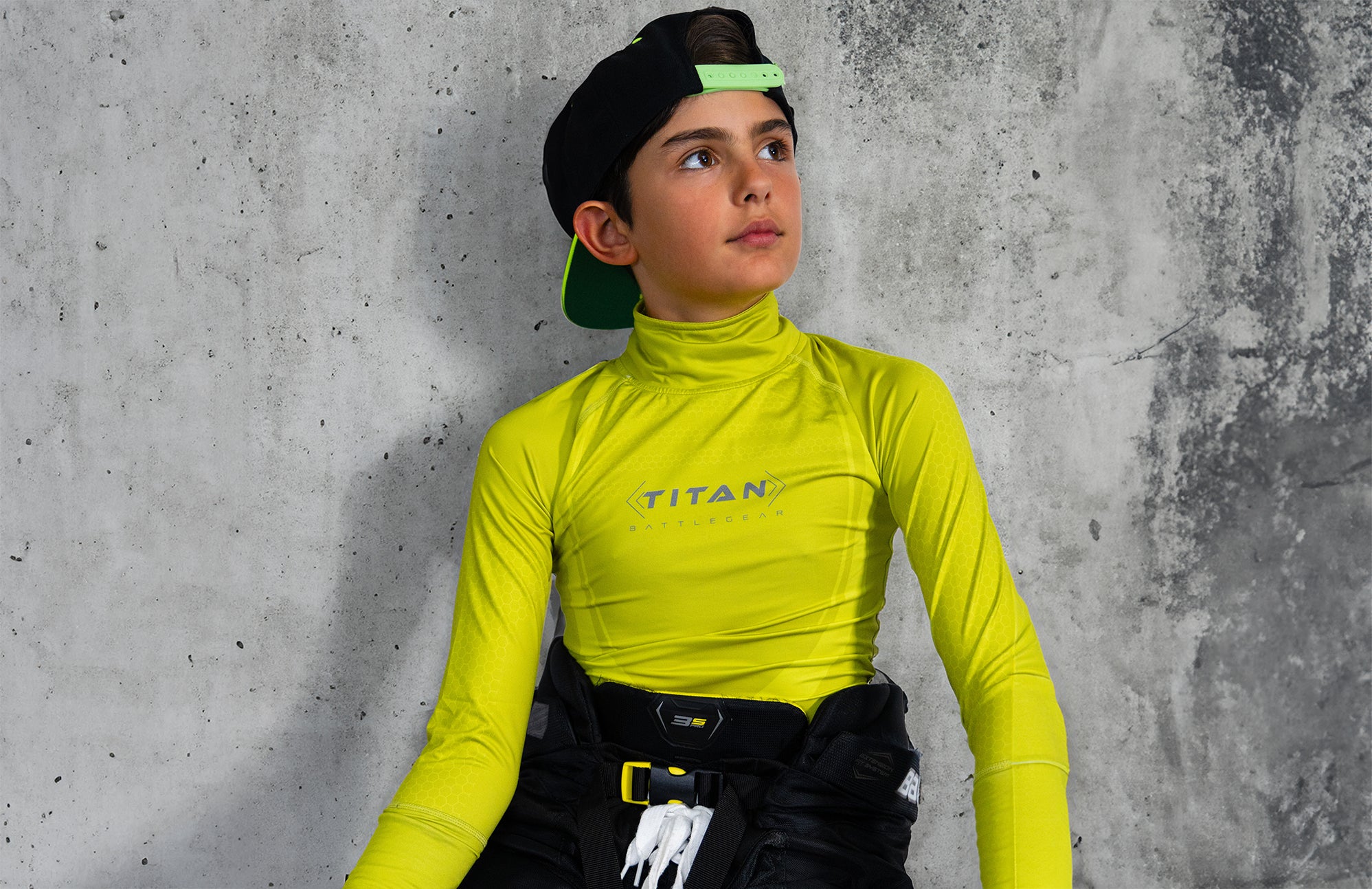 A youth hockey player wearing a bright yellow Titan BattleGear shirt with the Titan logo on the chest. The player is also wearing black hockey pants and a black cap with a green brim, sitting against a concrete wall.