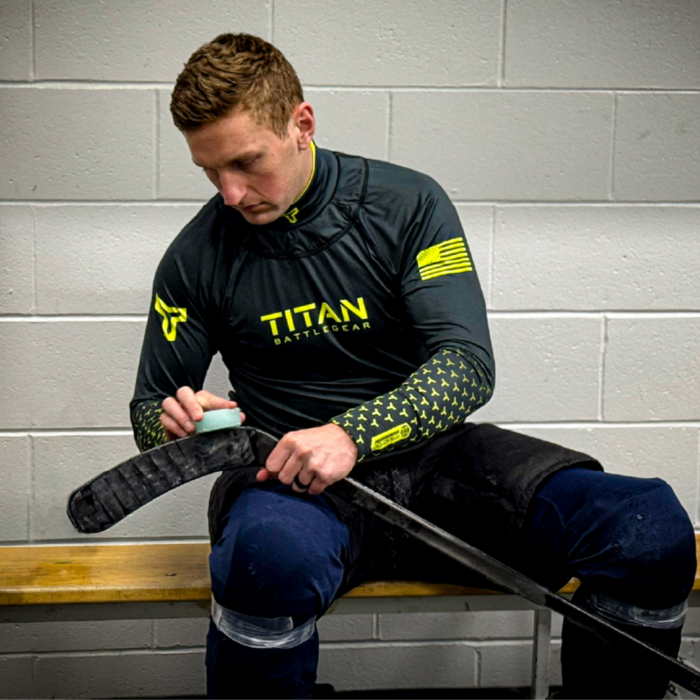 A hockey player sits on a locker room bench, taping his stick while wearing the Titan BattleGear 'Core Collection Flashpoint' long-sleeve shirt. This performance-driven base layer integrates advanced hockey neck guard technology, offering elite-level cut resistance and protection. The sleek black and neon yellow design reflects Titan’s commitment to both safety and bold aesthetics, ensuring players stay protected and confident on the ice.