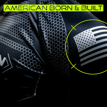 Close-up image of a Titan Battlegear shirt with an American flag emblem on the sleeve. The text "American Born & Built" is displayed at the top, highlighting the shirt's American manufacturing origin. The image emphasizes the quality and patriotic pride of the product.